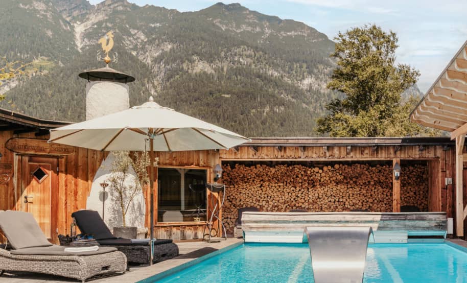 Outdoor pool in the Staudacherhof's wellness area with loungers and parasols, surrounded by an idyllic mountain landscape and a wood-burning sauna
