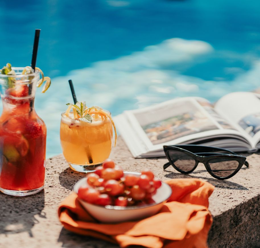 Erfrischende Cocktails und frische Früchte am Pool im Urlaub genießen Zwei Cocktails und eine Schüssel mit Trauben auf einem Steinrand, neben einem Buch und Sonnenbrille am Pool für einen entspannten Urlaub.