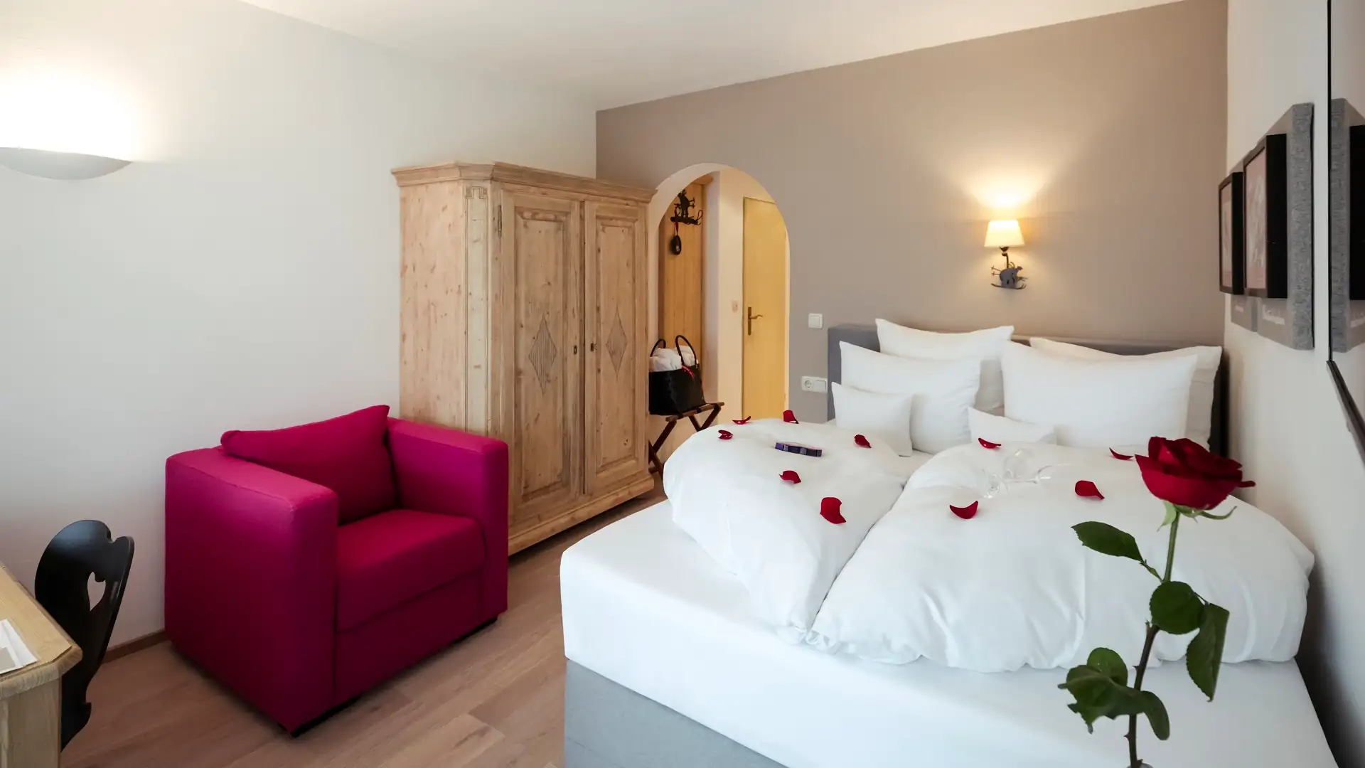 Romantisches Doppelzimmer in einem Hotel mit Rosenblättern auf dem Bett und einem pinken Sessel für einen perfekten Paarurlaub.