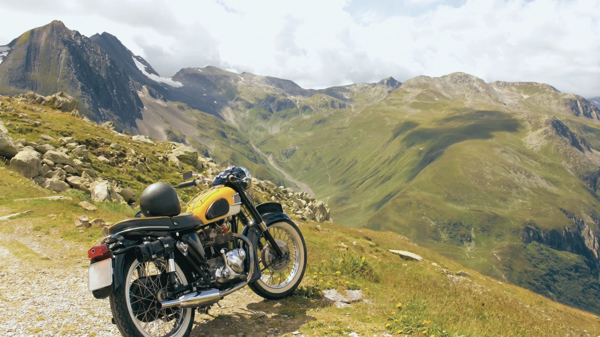 Ein klassisches Motorrad steht auf einem Bergweg in den Alpen, mit einer beeindruckenden Aussicht auf grüne Hügel und Berge.