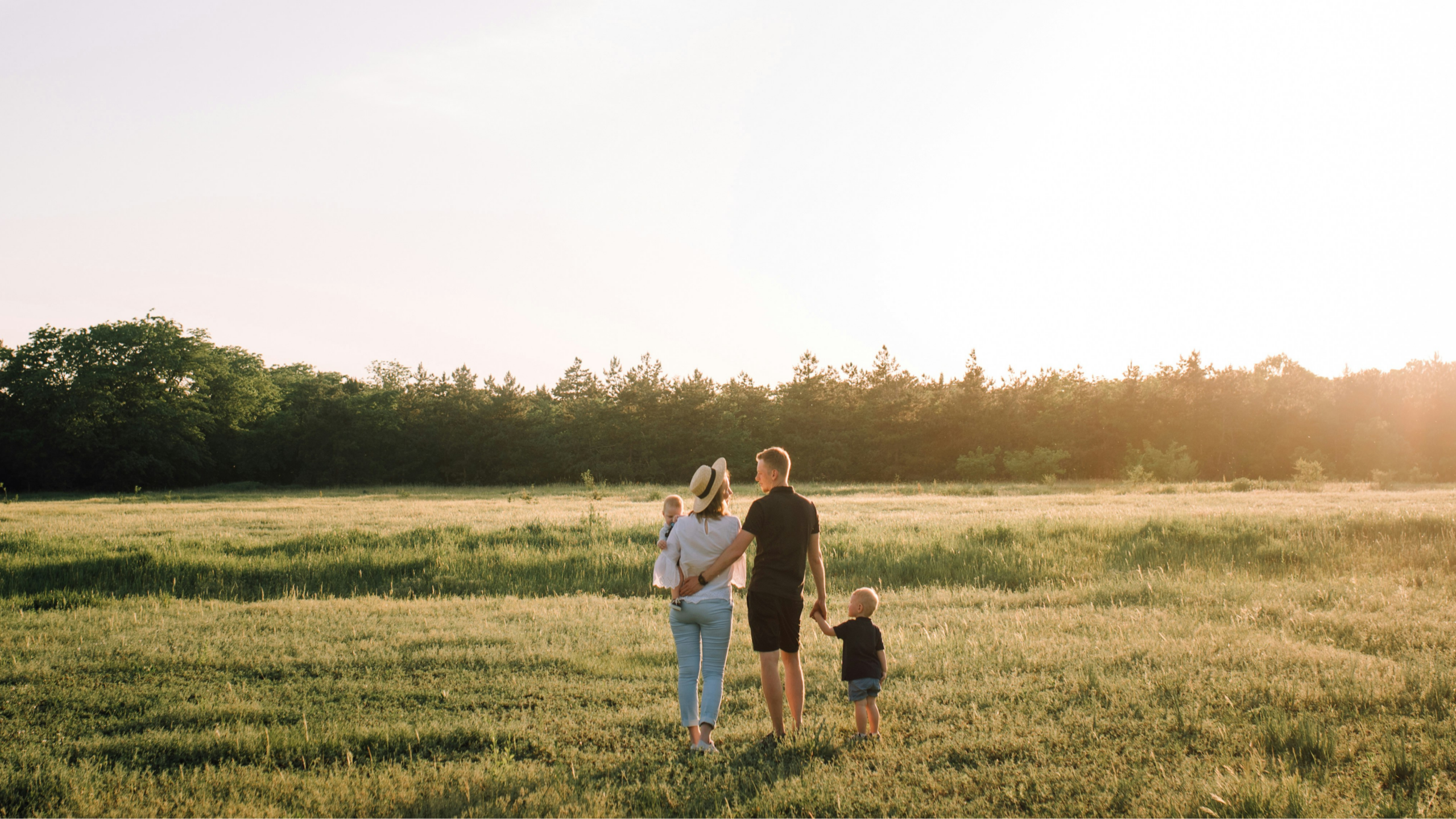 Eine Familie mit zwei kleinen Kindern spaziert auf einer Wiese bei Sonnenuntergang, umgeben von Bäumen.